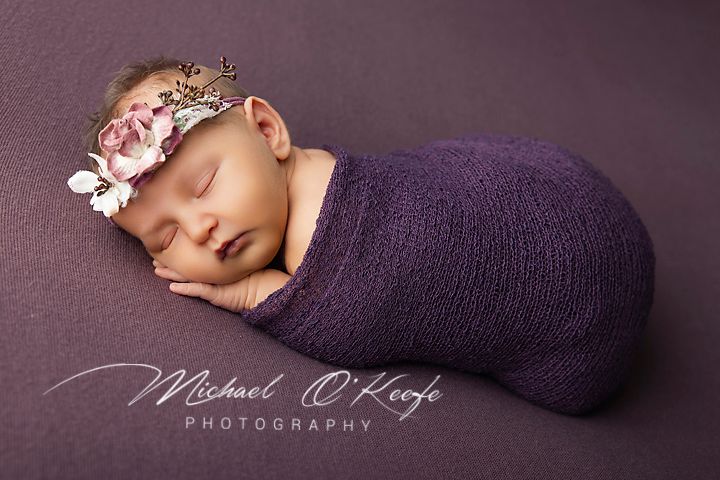 Newborn baby girl wrapped in purple swaddle with floral headband, sleeping peacefully on purple backdrop.