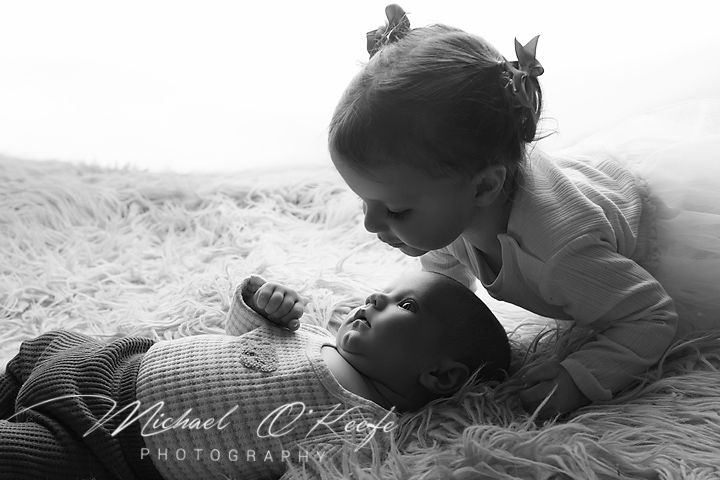 Black and white photo of a young girl tenderly kissing a newborn baby lying on a fluffy rug.