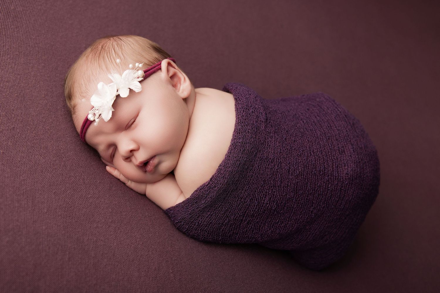 Sleeping newborn baby girl swaddled in purple wrap with floral headband on matching purple background.