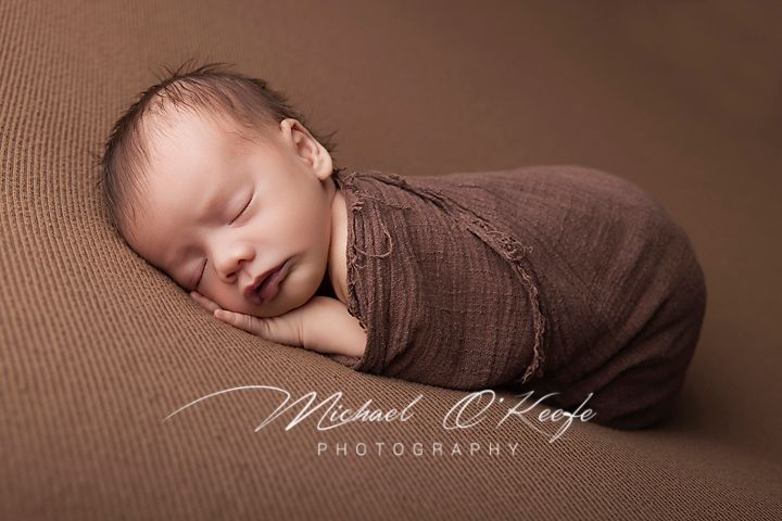 Peaceful newborn baby swaddled in brown wrap sleeping on matching brown backdrop, professional newborn photography.