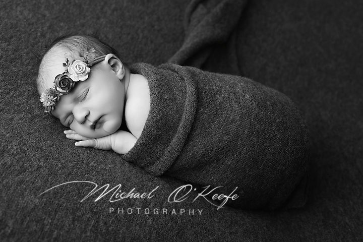 Black and white newborn photo of a swaddled baby girl with a floral headband, sleeping peacefully on a soft blanket.