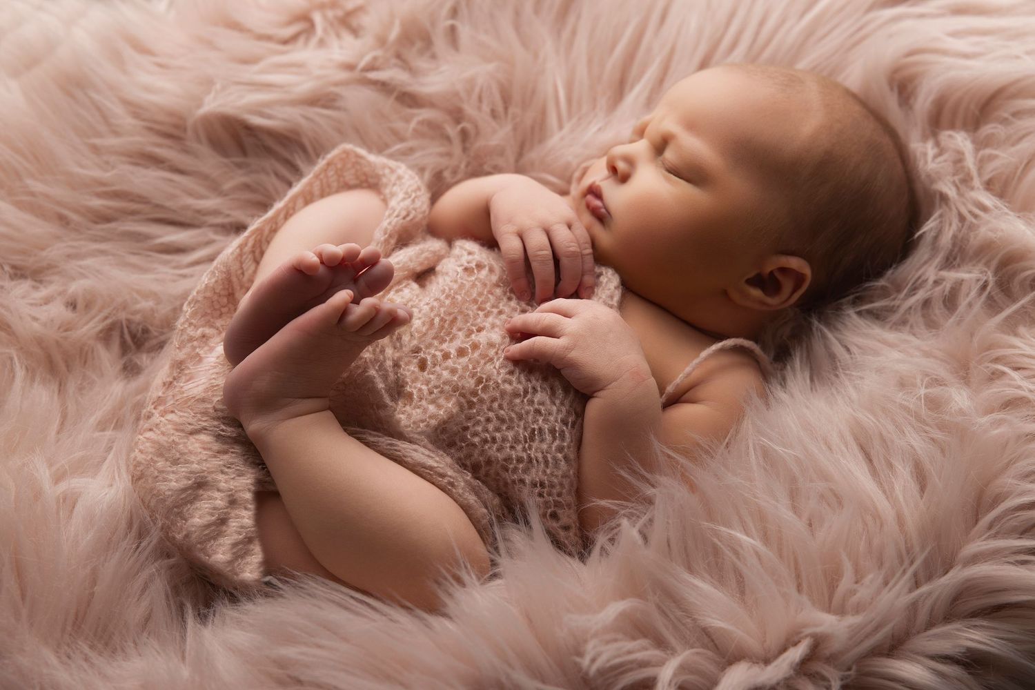 Peaceful sleeping newborn baby lying on soft pink fur blanket wearing knit wrap during professional photoshoot.