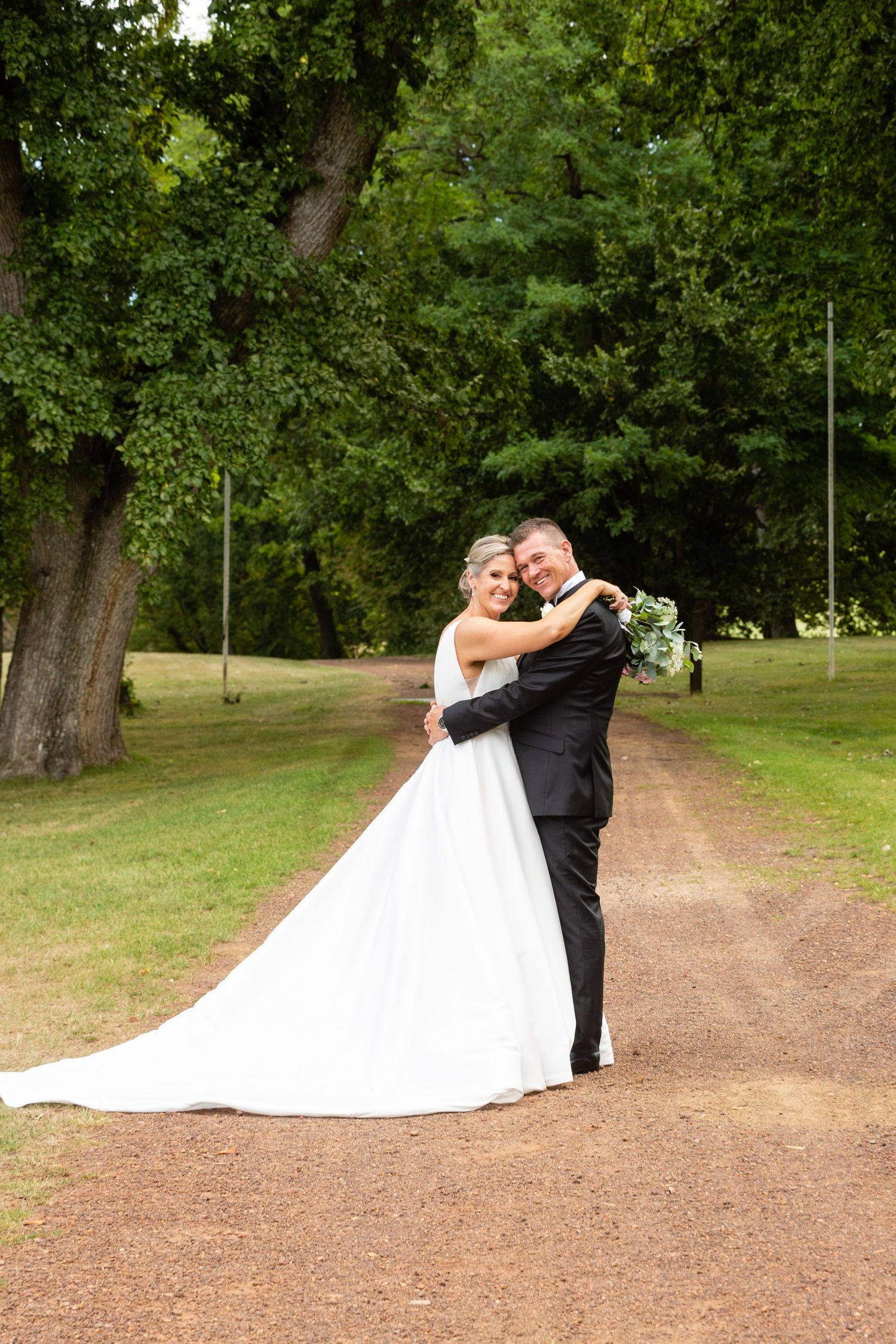 A romantic wedding portrait of a couple embracing on a tree-lined path in a park.