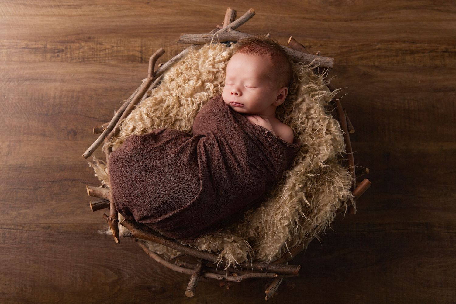 A brown newborn baby doll lies on rustic wooden surface with straw or hay bedding in multiple poses.