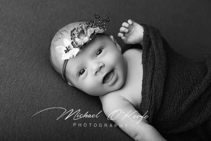 Black and white newborn photo of smiling baby wrapped in dark cloth with floral headband by Michael O'Keefe Photography.