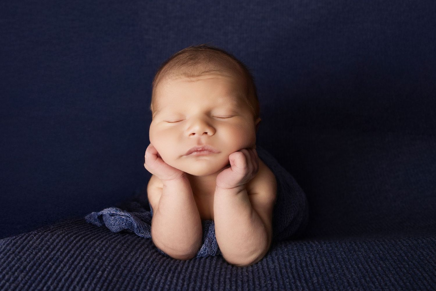 Newborn resting with hands under chin on navy blue textured backdrop.