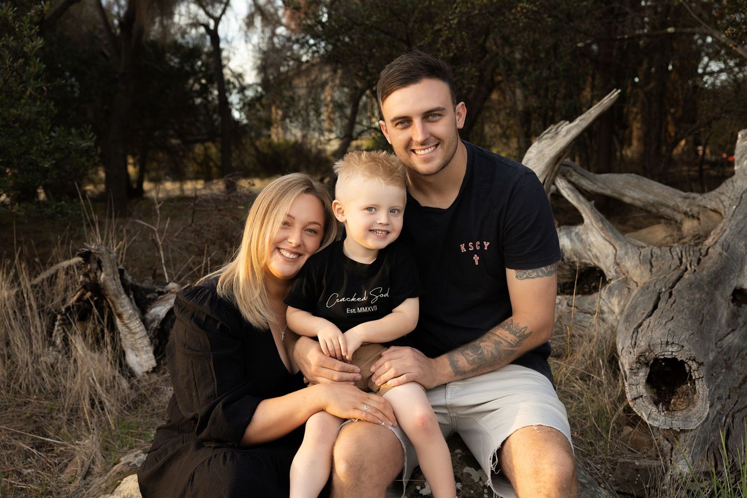 Family of three sitting together on rustic wooden log in outdoor setting.