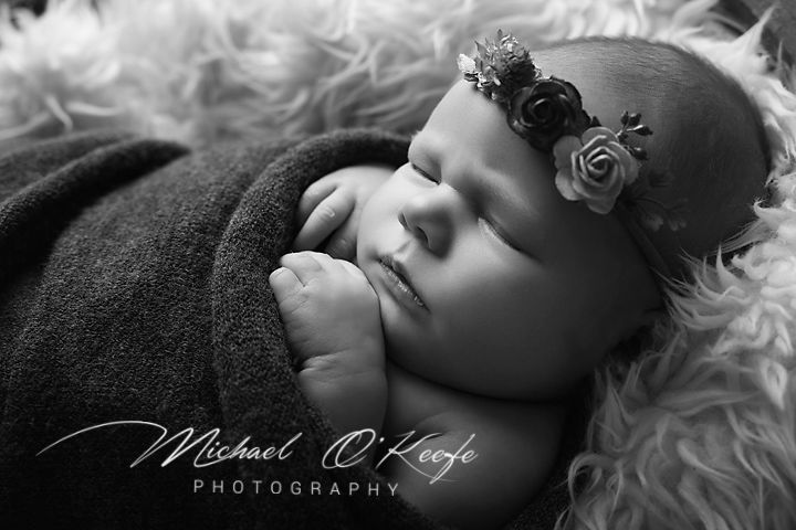 Black and white newborn photography of sleeping baby wrapped in dark blanket with floral headband by Michael O'Keefe.
