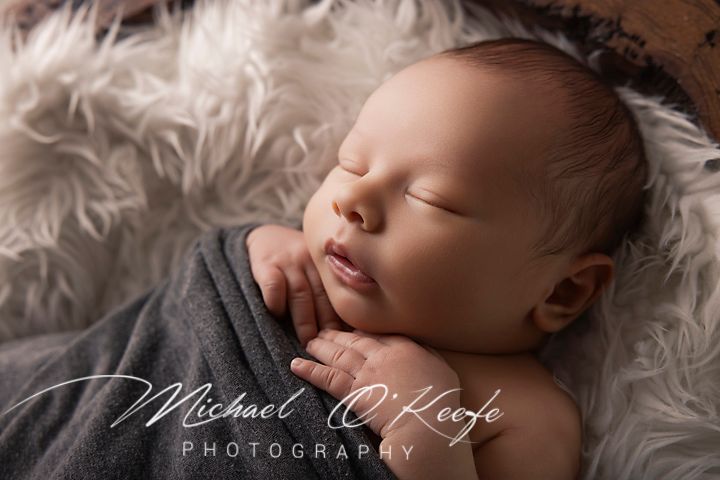Peaceful newborn baby sleeping wrapped in gray swaddle on fluffy white fur, professional newborn photography by Michael O'Keefe.