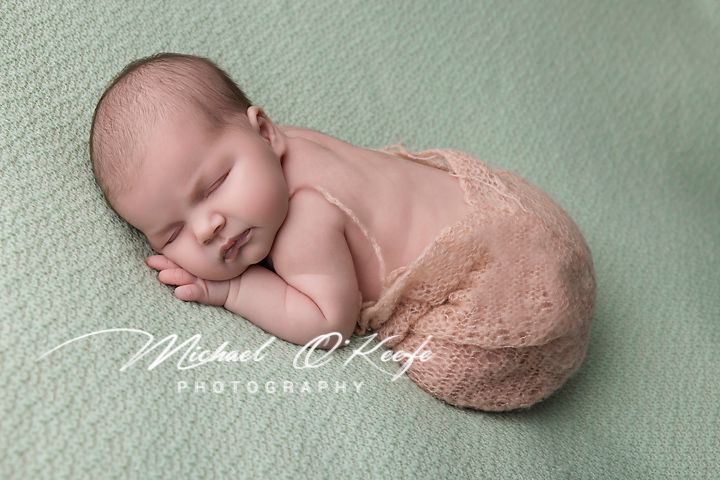 A sleeping newborn baby wrapped in pink knit blanket on sage green background.