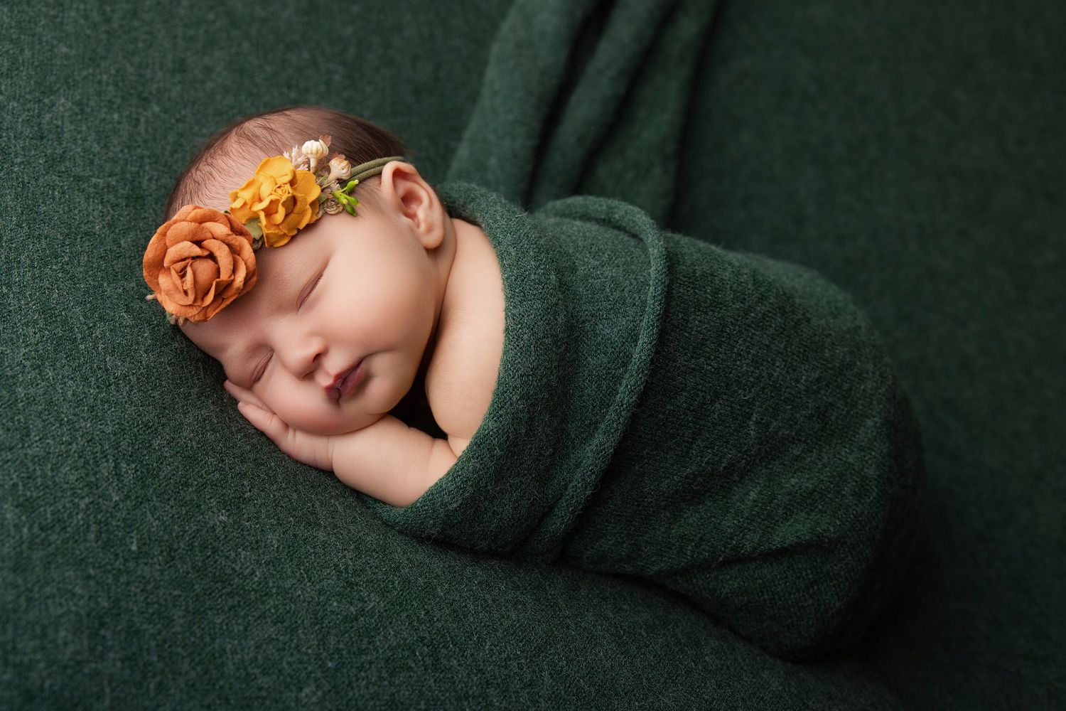 Newborn baby girl sleeping peacefully wrapped in green swaddle with orange floral headband on green background.