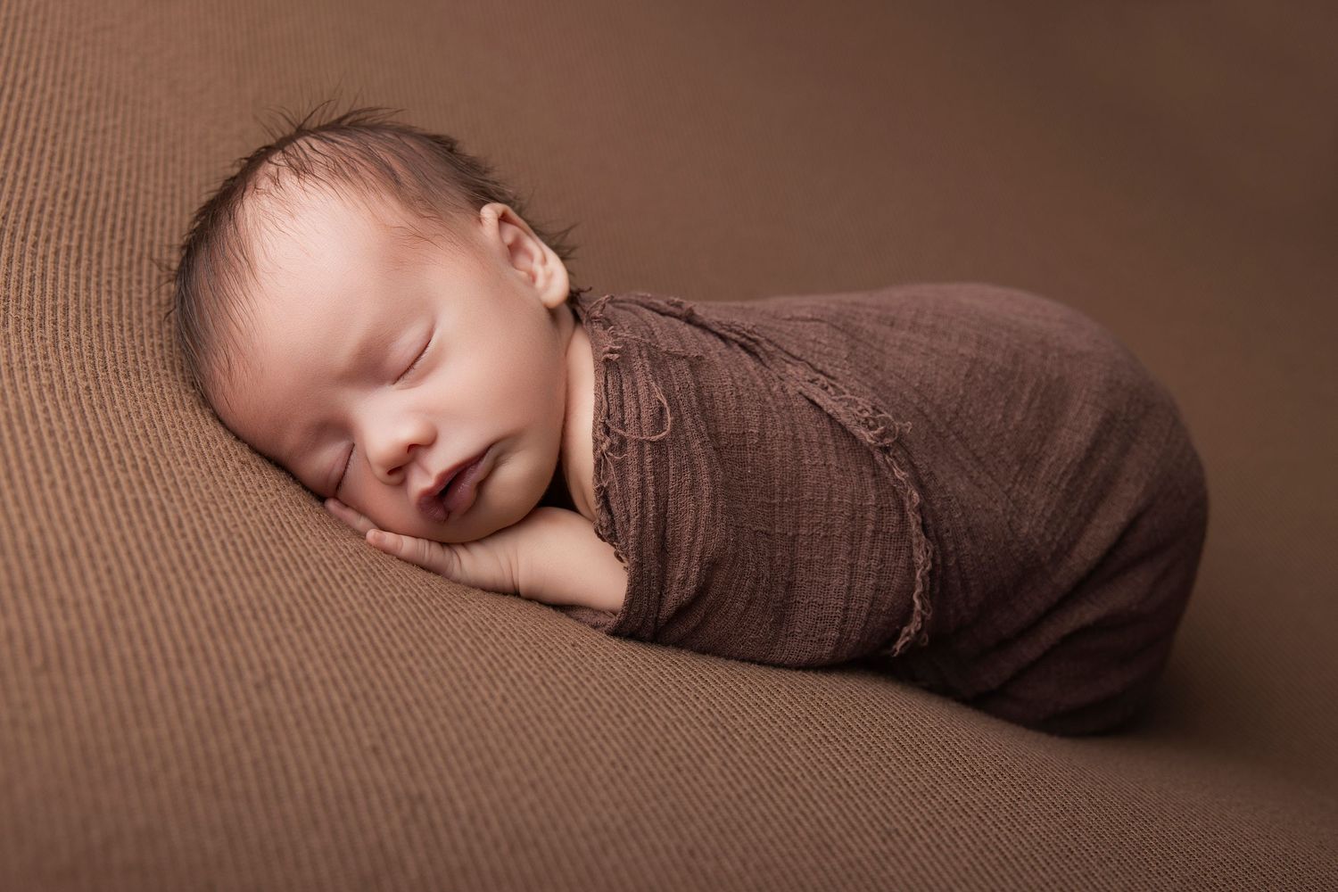Newborn baby swaddled in brown wrap sleeping peacefully on a matching brown textured background.