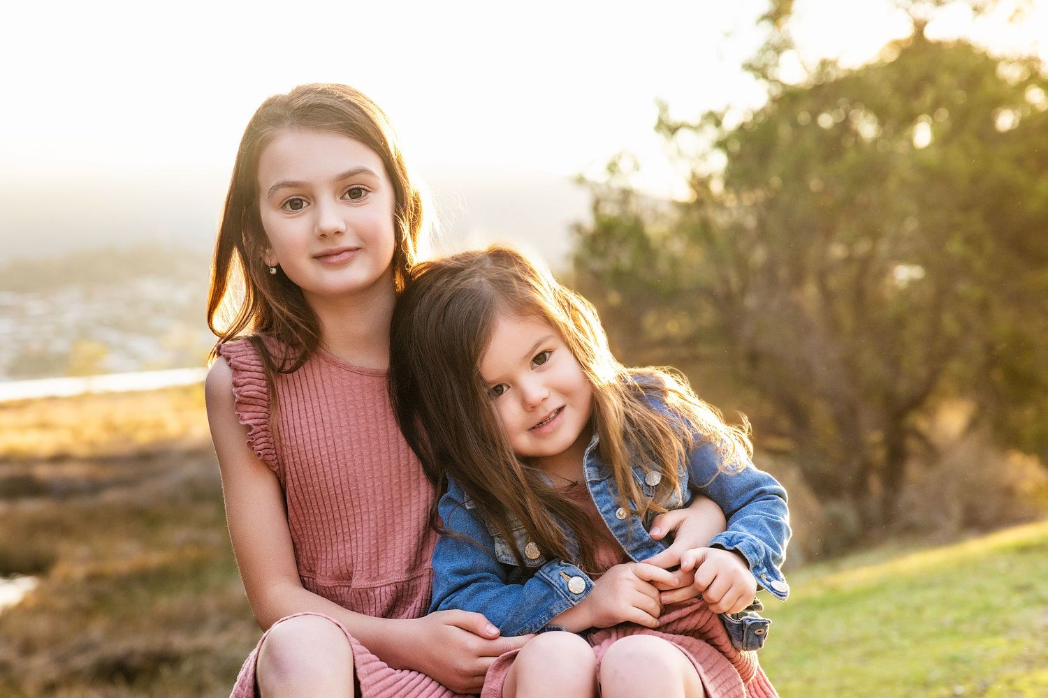 Two sisters sharing tender moment together at sunset on grassy hillside.