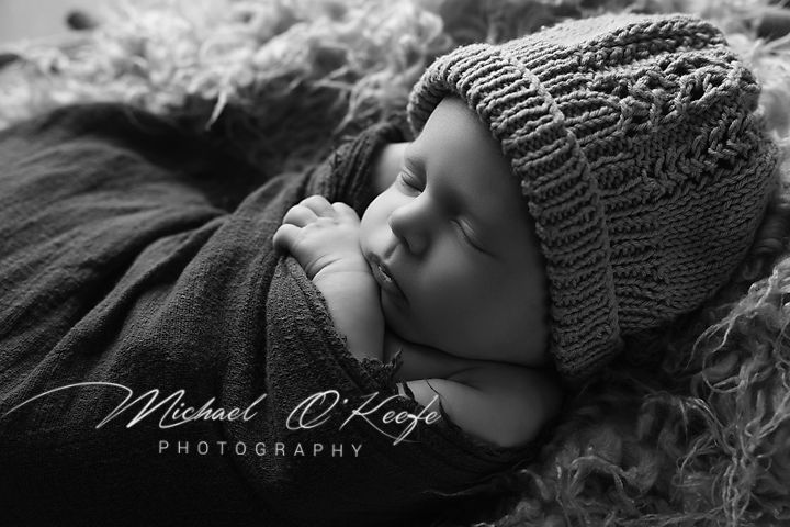 Black and white newborn photo of sleeping baby in knit hat, wrapped in soft blanket by Michael O'Keefe Photography.
