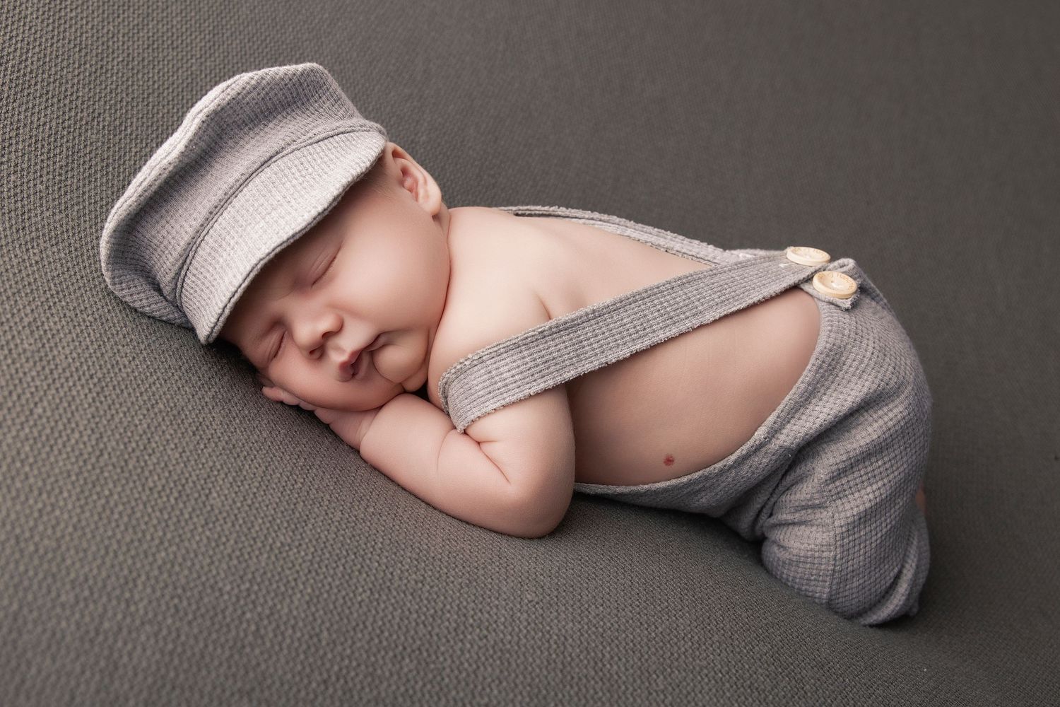 Sleeping newborn baby wearing gray knit overalls and matching flat cap posed on gray background.