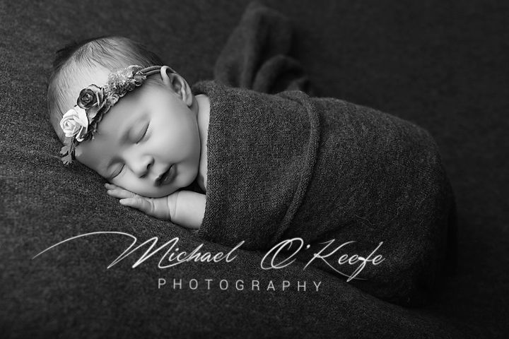 Black and white newborn photo of sleeping baby wrapped in gray swaddle with floral headband by Michael O'Keefe Photography.