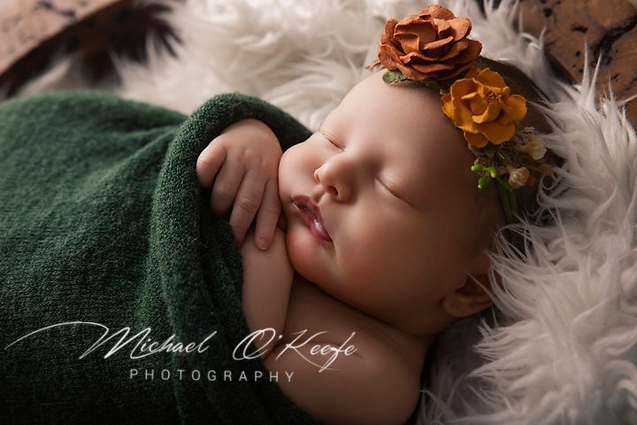 Newborn baby sleeping peacefully wrapped in green knit blanket with orange floral headband on fluffy white backdrop.
