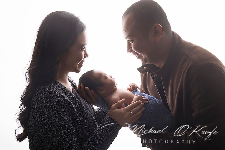 New parents lovingly gaze at their newborn baby in a professional studio portrait by Michael O'Keefe Photography.