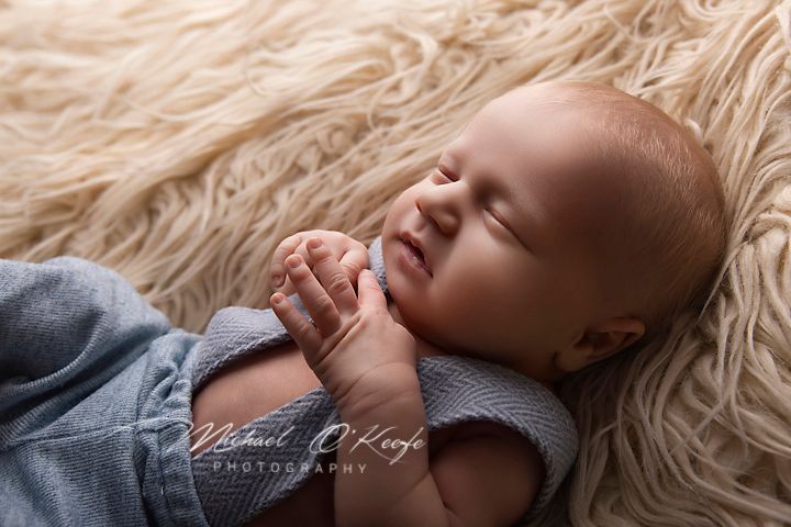 Sleeping newborn baby smiling with hands clasped, lying on a soft cream fur blanket in a blue wrap.