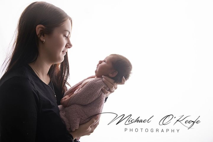 A mother tenderly holds her swaddled newborn baby in a soft, glowing studio portrait by Michael O'Keefe Photography.