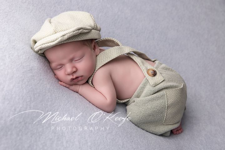 A sleeping newborn baby wearing a soft gray newsboy cap rests peacefully on a gray backdrop.