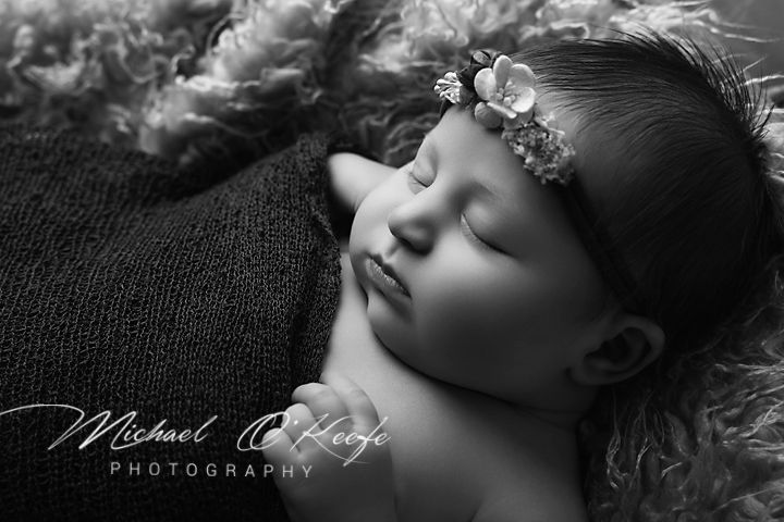 Black and white newborn photo of a sleeping baby girl with a floral headband resting on soft fluffy fur and a knit blanket.
