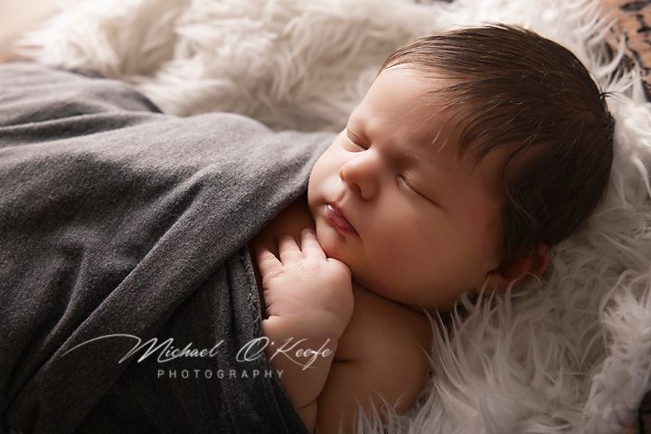 Sleeping newborn baby wrapped in gray blanket rests peacefully on white fur during professional photo session.