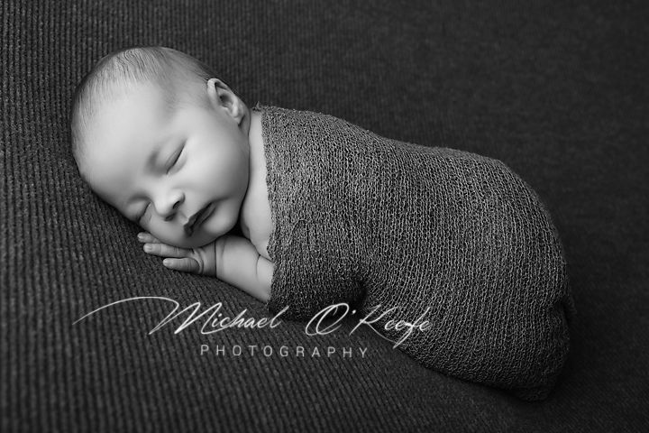 Black and white newborn photography of a sleeping baby swaddled in a gray wrap by Michael O'Keefe Photography.