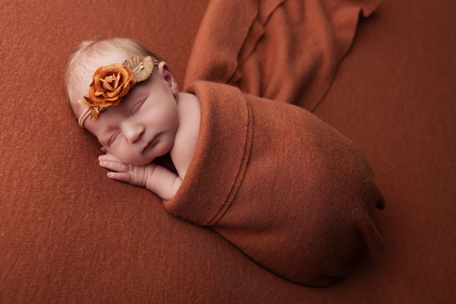 Sleeping newborn baby girl swaddled in rust orange wrap with floral headband on matching brown background.