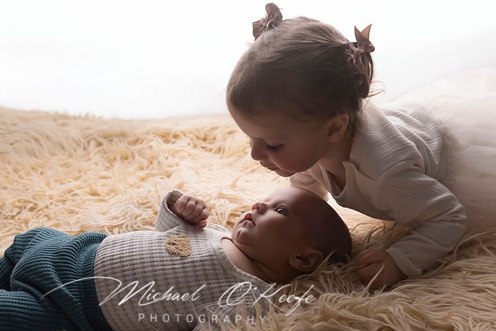 Young girl tenderly kisses newborn baby sibling lying on fluffy fur rug in soft natural light photography.