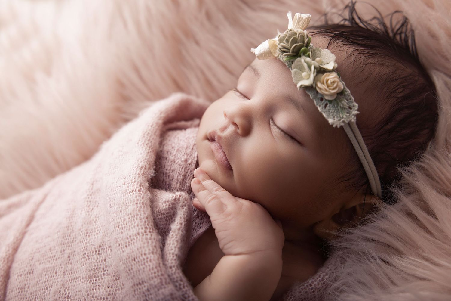 Peaceful newborn baby girl sleeping wrapped in pink, wearing a delicate floral headband on a soft fur backdrop.