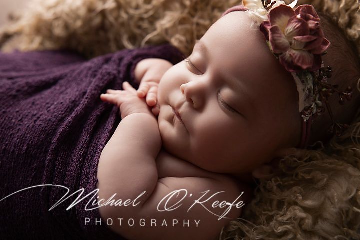 Newborn baby sleeping peacefully wrapped in purple, wearing floral headband, professional photography by Michael O'Keefe.