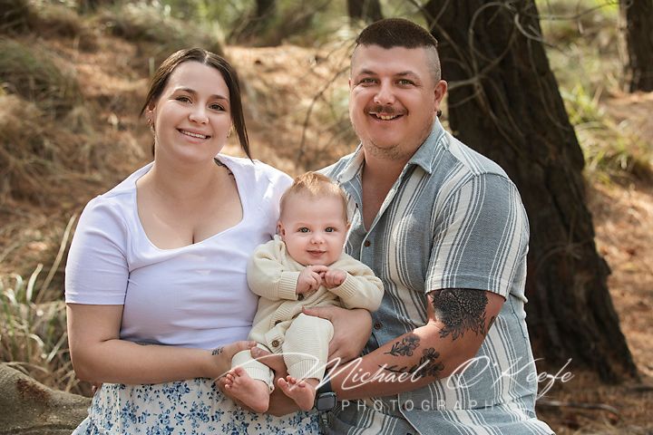 A family sits together outdoors on fallen logs in a wooded forest setting during a photo session.
