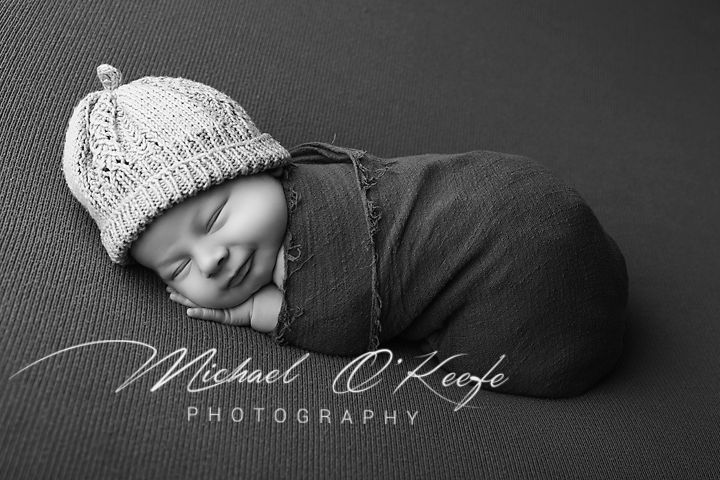 Black and white newborn photo of sleeping baby wrapped in dark swaddle wearing knit hat by Michael O'Keefe Photography.
