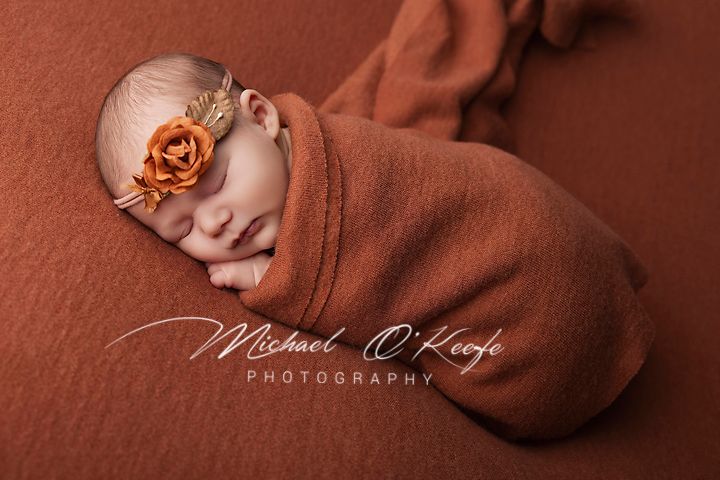 Newborn baby swaddled in rust orange wrap with floral headband, sleeping peacefully on matching background.