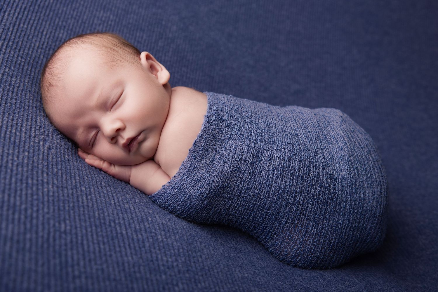 A sleeping newborn baby wrapped in a soft gray knit blanket against a dark blue background.