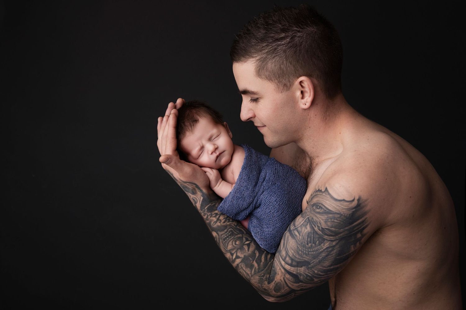 A tender newborn photography session featuring protective arms holding a sleeping baby against black background.
