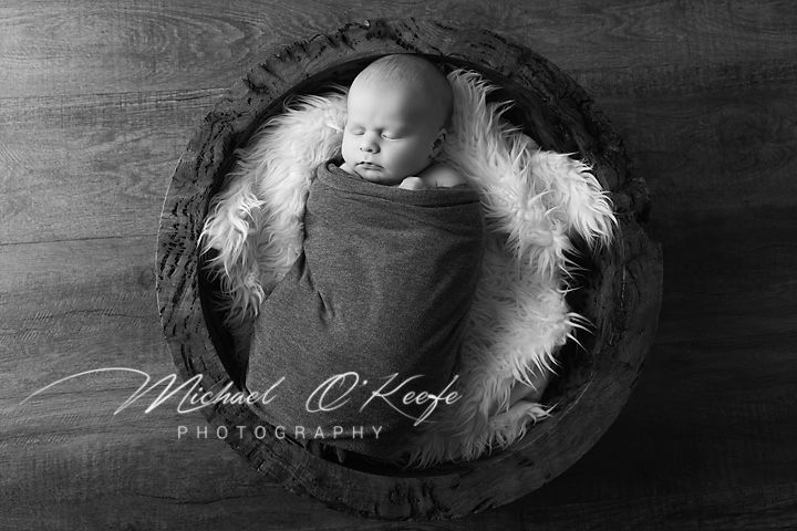 Moody newborn portrait in wooden bowl with dramatic lighting and watermark.
