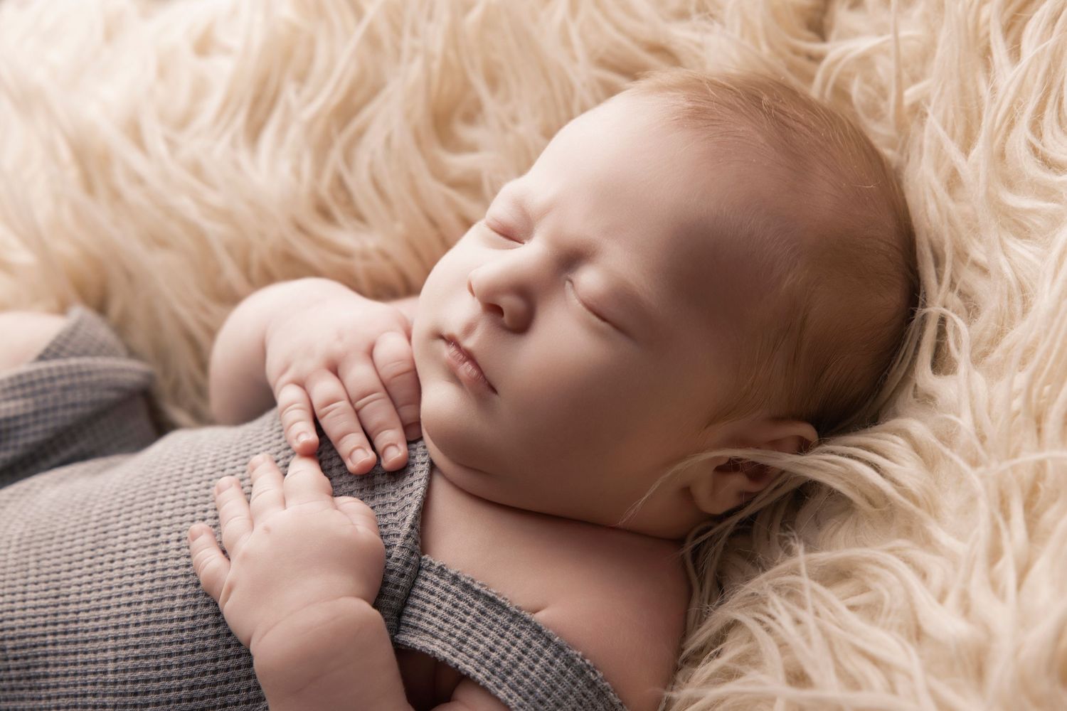 Sleeping infant in grey knit outfit on cream fur backdrop.