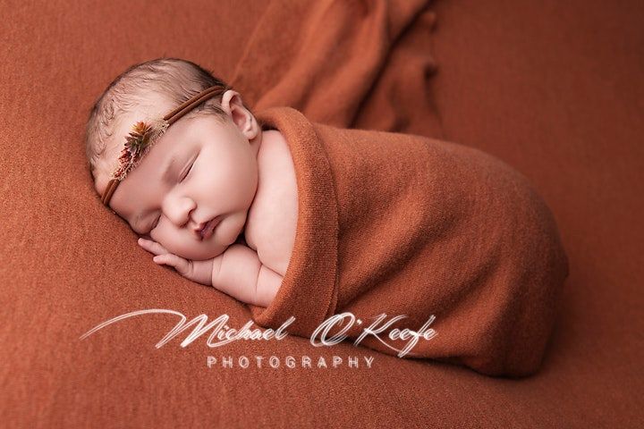 Sleeping newborn wrapped in orange fabric on matching backdrop.