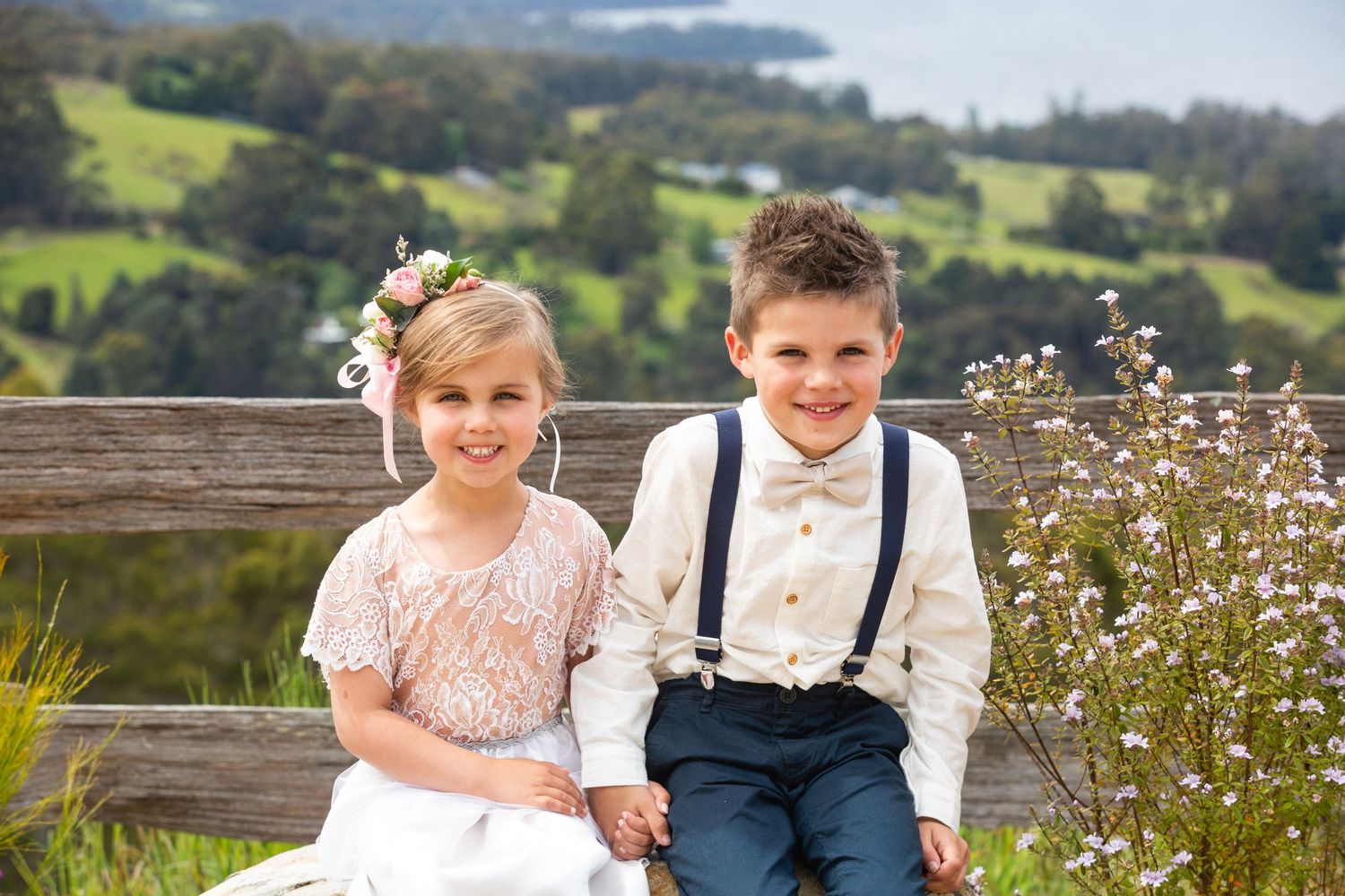Two young children in formal attire sit on a wooden bench against a rolling hillside backdrop.