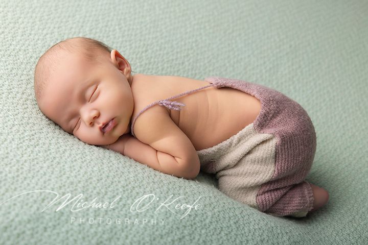 Newborn baby sleeping peacefully on mint green blanket wearing knit mauve overalls in professional photography studio.