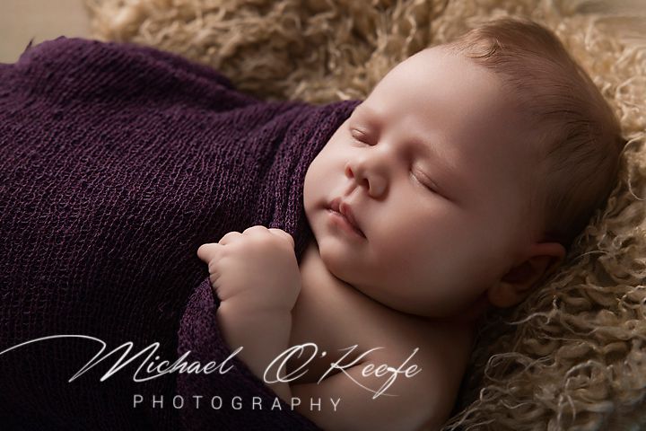 Newborn baby sleeping peacefully wrapped in a purple knit blanket on a fluffy beige backdrop by Michael O'Keefe Photography.