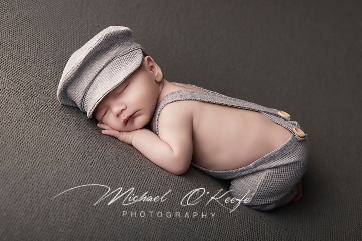 Newborn baby sleeping peacefully wearing striped newsboy cap and suspenders outfit on gray background.