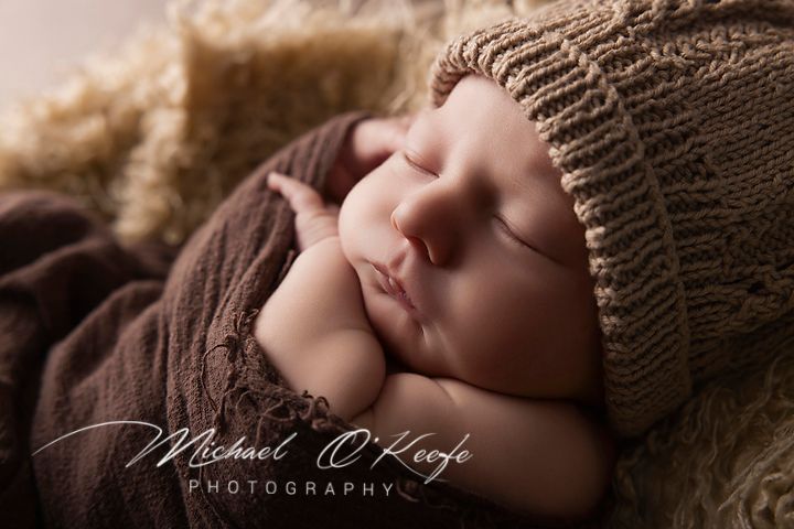 Peaceful newborn baby sleeping in brown knit hat and wrap, professional newborn photography by Michael O'Keefe.