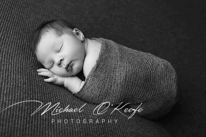 Black and white newborn photography session showing baby wrapped in soft fabric against dark textured background.