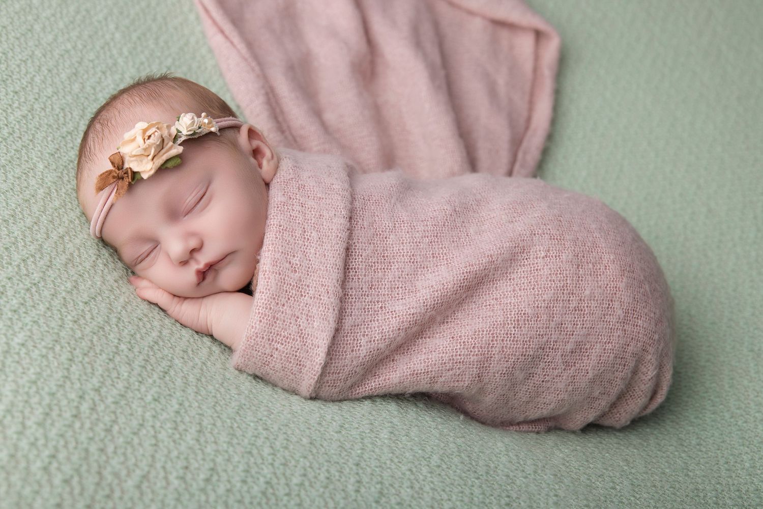 Sleeping newborn baby swaddled in pink wrap with floral headband on mint green background.
