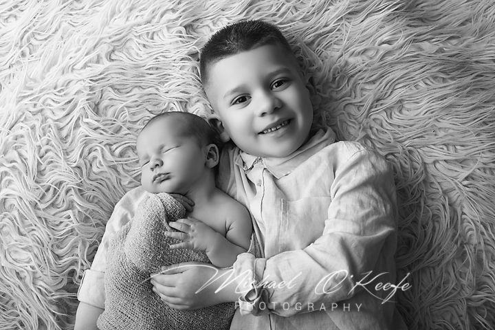 Black and white photo of a smiling boy holding a sleeping newborn baby on a fluffy white blanket.