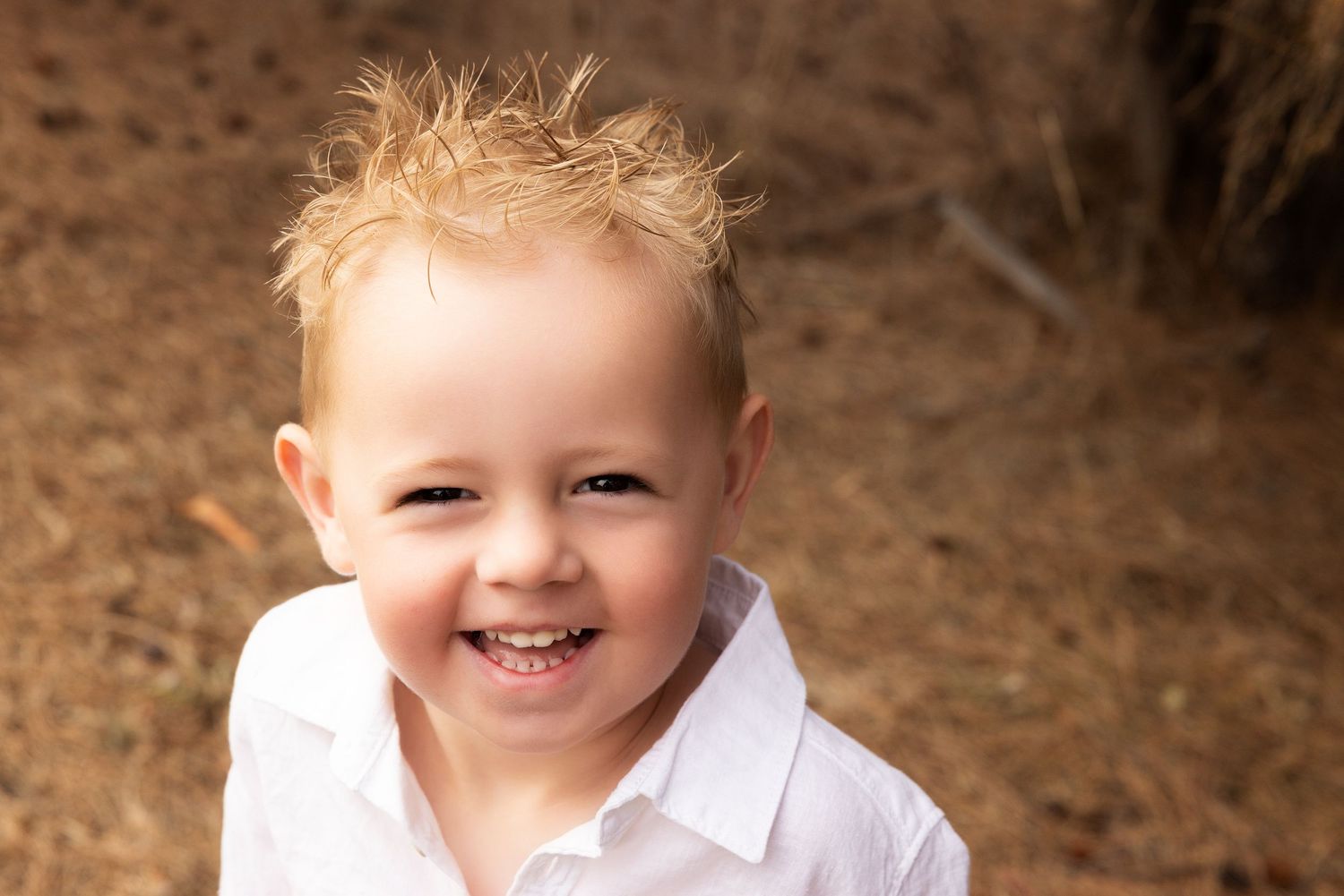 Close-up portrait of a smiling child with blonde spiky hair wearing white shirt outdoors.
