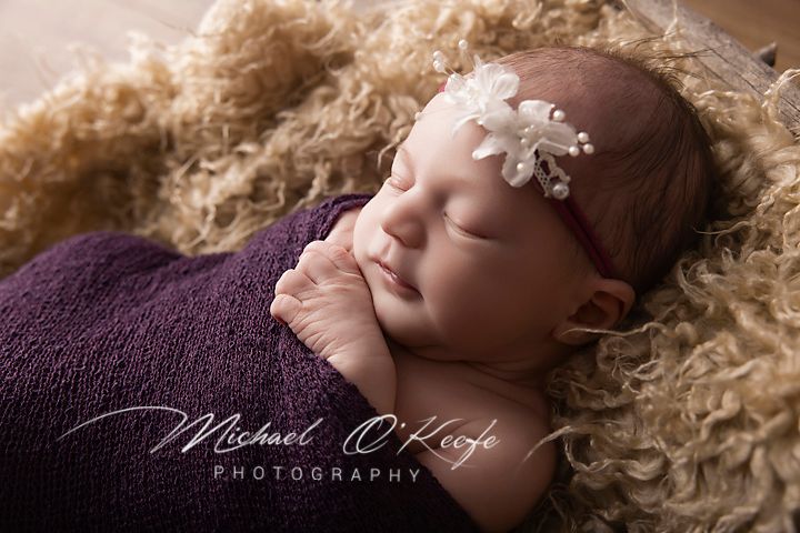 Newborn baby girl sleeping peacefully wrapped in purple, wearing a white flower headband on a fluffy tan blanket.